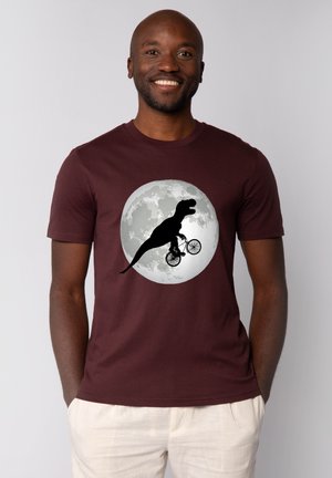 Smiling man wearing a maroon t-shirt with a graphic of a T-Rex riding a bicycle in front of a full moon.