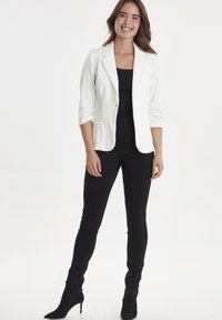 Blazer bianco con vestibilità su misura, chiusura a un bottone e maniche arrotolate, abbinato a un top nero e pantaloni neri slim-fit, indossando scarpe col tacco nere.