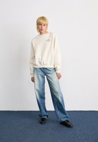 Persoon met kort blond haar, gekleed in een crèmekleurige sweatshirt, lichtblauwe wijde jeans en zwarte schoenen, staat op een blauwe vloerbedekking.