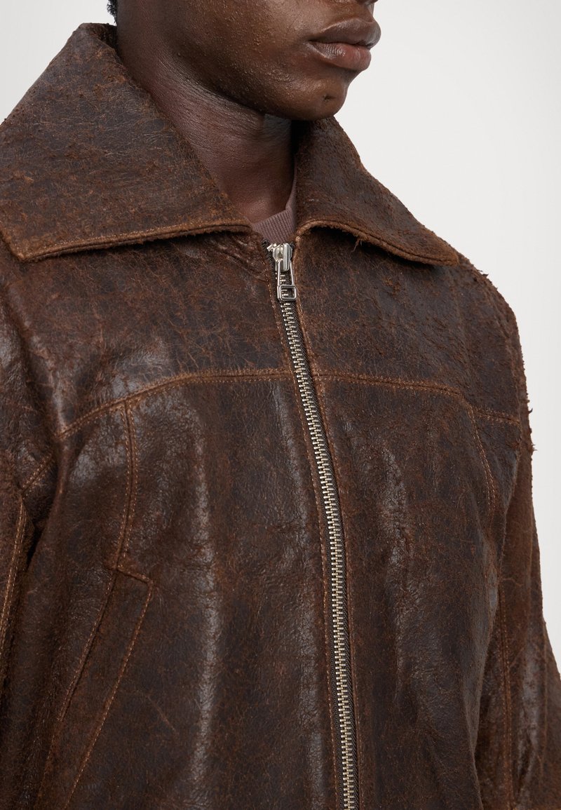 Brown leather jacket featuring a structured collar, zip closure, and textured surface. Detailed seam lines and side pockets visible.