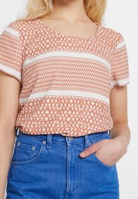 Camiseta de manga corta en melocotón con patrones y rayas geométricas blancas, hecha de tejido ligero, combinada con jeans de mezclilla azul de talle alto.