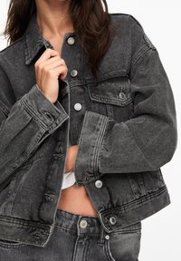 Chaqueta de mezclilla negra con botones plateados, mangas anchas y diseño corto. Presenta una tela texturizada y bolsillos frontales.