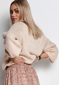 Jasno-beżowy, oversize'owy sweter z dzianiny o szerokich rękawach, w połączeniu z wzorzystą różową spódnicą z falami i subtelnymi akcentami kolorystycznymi.