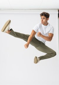 Hvid kortærmet t-shirt, olivengrønne træningsbukser og beige sneakers. Motivet udfører et højt spark og viser dynamisk bevægelse.
