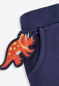 Pantaloni de trening bleumarin dintr-un material texturat, având o aplicație cu un dinozaur roșu și portocaliu, cu pete galbene în zona buzunarului.