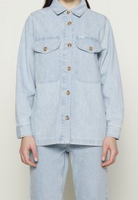 Niebieska kurtka dżinsowa z dwoma kieszeniami na piersi, złotymi guzikami i zapinana na guziki z przodu. Posiada klasyczny kołnierz oraz luźny krój.