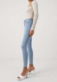 Jeans ajustados de color azul claro con una textura suave, de alta cintura y con un ligero estiramiento, combinados con tacones de bloque abiertos blancos y una blusa de punto beige.