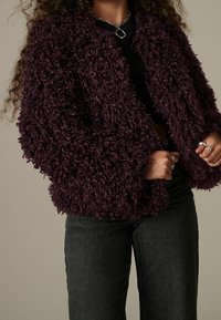 Donna che indossa una giacca in pelliccia sintetica riccia di colore viola scuro sopra un top nero e jeans scuri, con anelli d'argento e una collana a catena spessa.