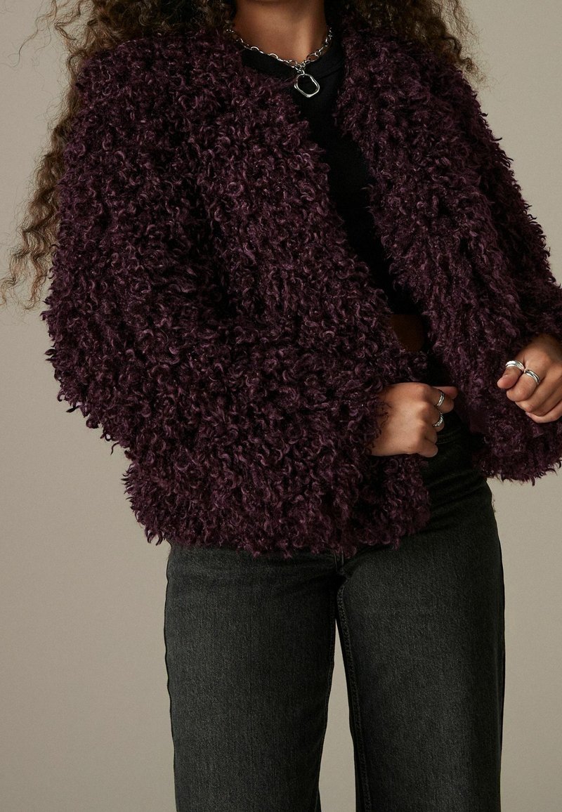 Donna che indossa una giacca in pelliccia sintetica riccia di colore viola scuro sopra un top nero e jeans scuri, con anelli d'argento e una collana a catena spessa.