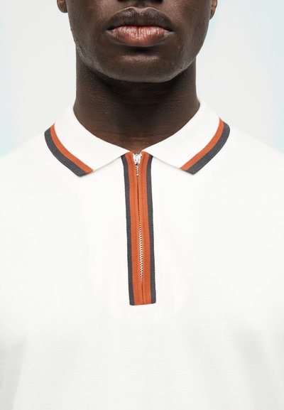 Κοντινό πλάνο ενός ατόμου που φορεί ένα λευκό πόλο πουκάμισο με πορτοκαλί και μαύρη ριγέ γιακά και φερμουάρ στο μπροστινό μέρος.