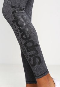 Svarta leggins med en melerad textur, prydda med en glänsande "Superdry"-logotyp på sidan. Tillverkade av elastiskt material för komfort och passform.