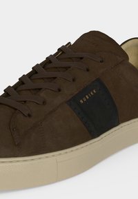Basket en daim brun foncé avec des lacets assortis, une semelle beige et une bande latérale noire portant l’inscription "NUBIKK" en lettres dorées.