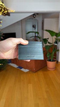 Musta nahkainen korttikotelo, jossa on sileä pinta, useita korttipaikkoja ja kohokuvioitu brändi edessä. Kädessä sisätilataustaa vasten.