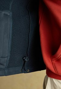 Jūras spēku zīmola flīsa jaka ar regulējamu apakšmali un zelta detaļām, apvienota ar sarkanu sporta krekls, kas izceļas ar gludu tekstūru un kapuci.