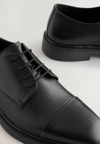Zapatos de cuero negro con cordones, acabado liso, puntera redondeada y detalle de costura contrastante a lo largo de la suela y el borde del zapato.