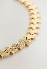 Kuldne tähtkujuline käevõru, millel on mõned tähed, mis on kaunistatud väikeste selgete rhinestone'idega heledal taustal.