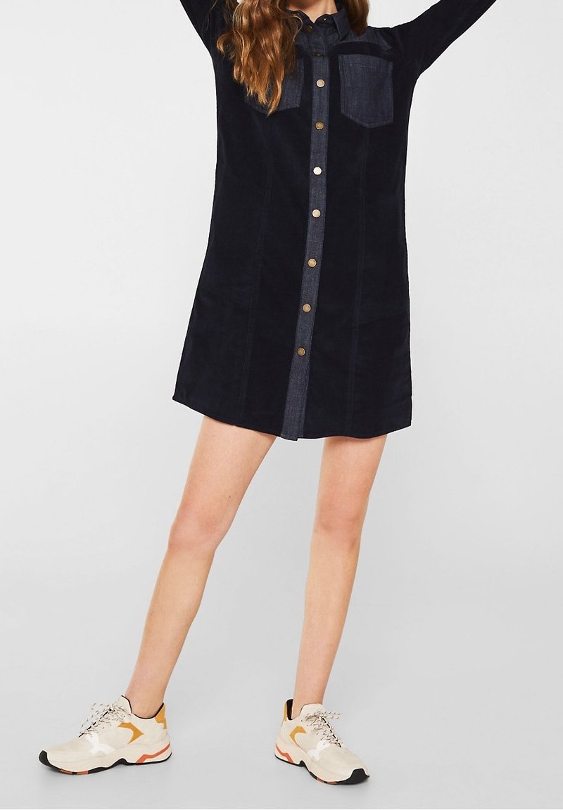 Femme portant une robe noire boutonnée avec une poche poitrine et des baskets beiges, debout jambes écartées et bras levés.
