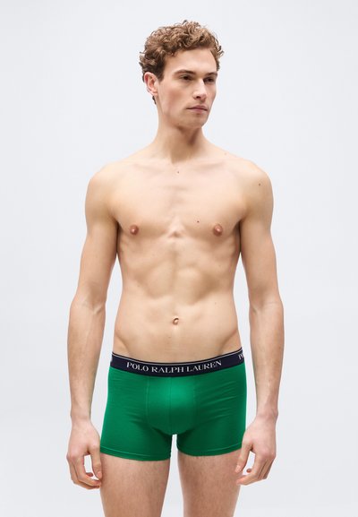 Νέος άνδρας με κατσαρά μαλλιά που φοράει πράσινα μποξεράκια Polo Ralph Lauren, στέκεται μπροστά σε απλό φωτεινό γκρι φόντο.