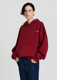 Felpa con cappuccio color mogano con tasca frontale e testo ricamato. Tessuto morbido, vestibilità oversize, abbinata a jeans blu scuro.