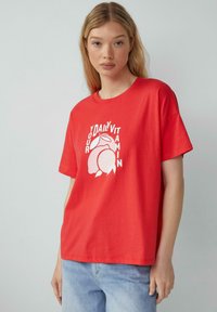 Rode katoenen t-shirt met een ronde hals en korte mouwen, voorzien van een witte grafische afbeelding met fruit en de tekst "Je Dagelijkse Vitamine."