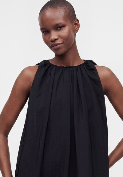 Femme aux cheveux courts portant une robe noire ample sans manches avec un col froncé, debout devant un fond clair uni.