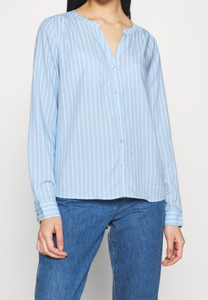 Vrouw draagt een lichtblauw, lang mouw, verticaal gestreept bloesje met knoopjes, gecombineerd met blauwe jeans.