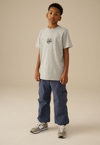 Maglietta di cotone grigia con logo rotondo, pantaloni cargo blu navy con cerniere laterali e sneakers grigie con accenti chiari. Completo semplice e casual.