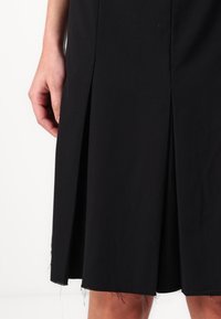 Fustă neagră cu un design plisat și detalii cu margine brută, realizată dintr-un material neted. Mână în cadru, arătând lungimea îmbrăcămintei.