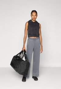 Felpa nera corta, pantaloni della tuta grigi e sneakers nere. Tiene una grande borsa da viaggio nera. Tessuto morbido, vestibilità rilassata, dettagli minimi.