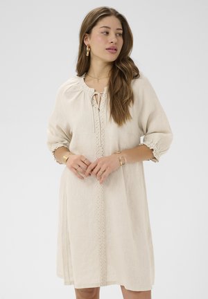 Žena s dugom smeđom kosom koja nosi bež haljinu dužine do koljena s vezenim prednjim dijelom, rukavima tri četvrtine i zlatnim nakitom, gleda udesno.