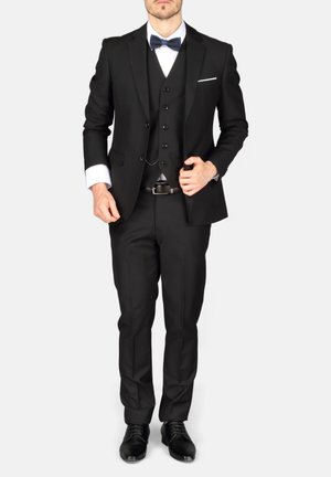 Homme portant un costume trois pièces noir avec une chemise blanche, un nœud papillon noir et des chaussures habillées noires sur un fond clair uni.