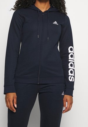 Person iført en sort Adidas lynlås-hættetrøje med hætte og hvidt logo på bryst og ærme, kombineret med matchende sorte bukser.