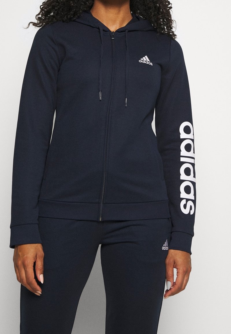 Personne portant un sweat à capuche zippé noir Adidas avec un logo blanc sur la poitrine et la manche, associé à un pantalon noir assorti.