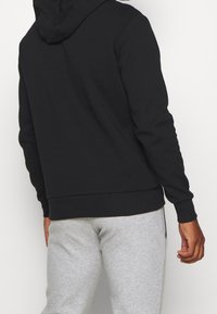 Sweat à capuche noir en tissu doux avec une poche kangourou et une capuche à cordon ajustable, porté avec un pantalon de survêtement gris clair.