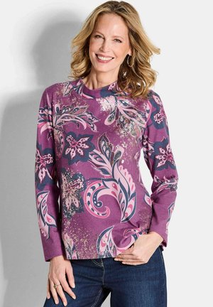 Lächelnde Frau mit blonden Haaren, die ein lila Langarmoberteil mit pinken und marineblauen Blumenmustern trägt, kombiniert mit dunkelblauen Jeans, steht vor einer weißen Wand.