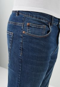 Jeans de mezclilla azul oscuro con un ajuste estándar, cinco bolsillos y herrajes de tono cobre con detalles de costuras visibles a lo largo de los costados.