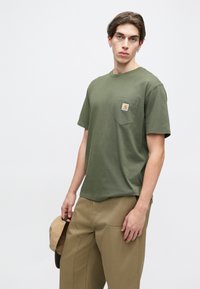 Olivově zelené bavlněné tričko s kulatým výstřihem a krátkými rukávy, s náprsní kapsou a logem. Volný střih a jemná textura.
