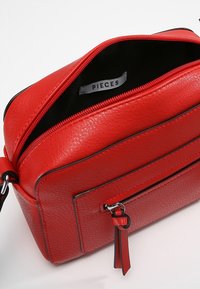 Červená kožená kabelka s texturovaným povrchem, zipovým uzávěrem, přední kapsou a černým interiérem. Obsahuje přívěsek s sítotiskem "PIECES".