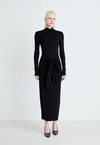 Μακριά μαύρη φόρεμα με μακριά μανίκια, κατεβασμένη μέση με διακοσμητικό κόμπο και λεία υφή. Φοριέται με μαύρα στρας σουέτ παπούτσια με τακούνι.