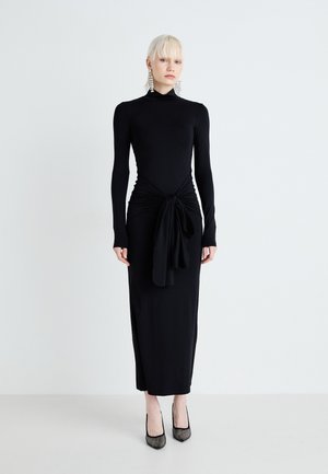 Long black dress with long sleeves, gathered waist featuring a knotted accent, and a smooth texture. Worn with black embellished heels.