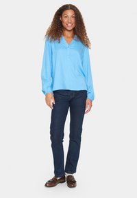 Blusa de manga larga azul claro con escote en V y cierre de botones, combinada con vaqueros de denim oscuro y zapatos marrones. Textura suave y corte relajado.
