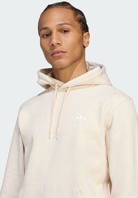 Felpa crema con cappuccio regolabile e tasca a marsupio; presenta un piccolo logo bianco Adidas sul lato sinistro del petto. Tessuto dalla texture liscia.