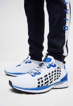 White and blue athletic sneakers with black checkered pattern worn with navy blue pants featuring blue text on the side.