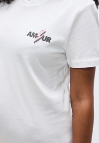 Άτομο που φοράει λευκό T-shirt με το κείμενο «AMOUR», όπου το γράμμα «O» αντικαθίσταται από ροζ κεραυνό στην περιοχή του στήθους.