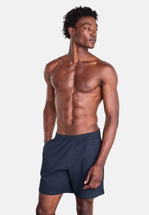Muskelbepackter Mann mit dunklem lockigem Haar, trägt marineblaue Shorts, steht mit den Händen in den Taschen und blickt nach links vor einem einfarbigen Hintergrund.