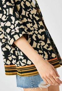 Frau trägt ein gemustertes schwarz-beige Shirt mit orangen Streifen und zerrissene blaue Jeansshorts, zeigt eine Hand mit einem goldenen Ring.