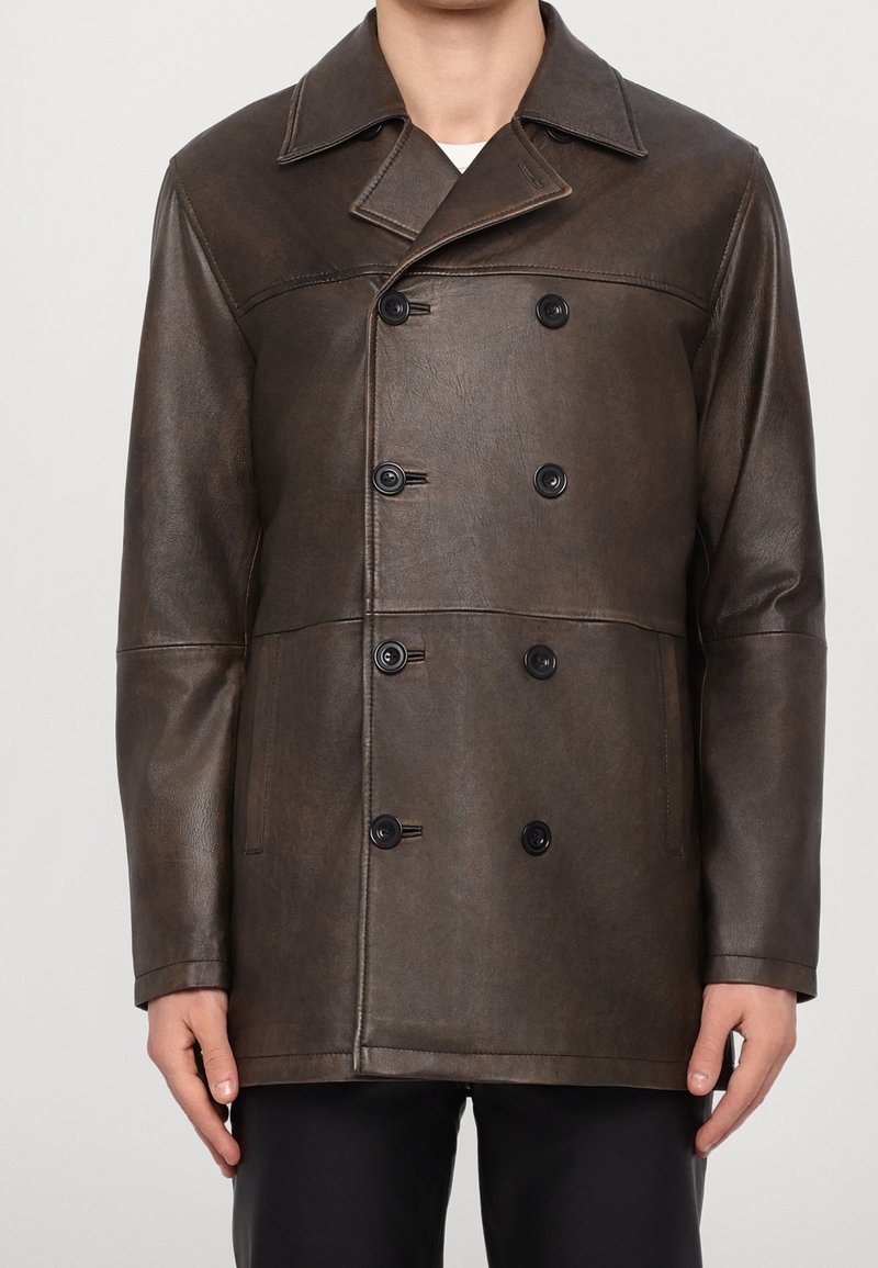 Veste en cuir marron foncé croisée avec des boutons noirs portée par une personne les mains détendues sur les côtés.