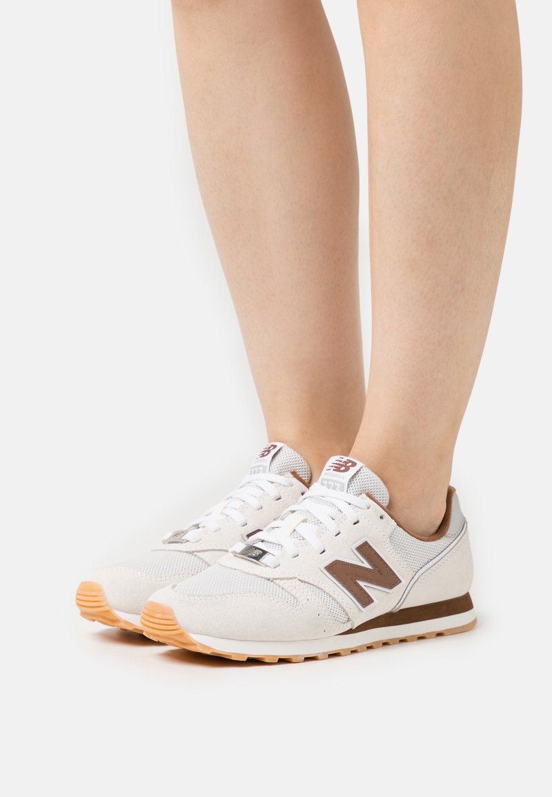 Jambes portant des baskets New Balance blanches et marron avec des lacets blancs et des semelles couleur gomme sur un fond blanc uni.