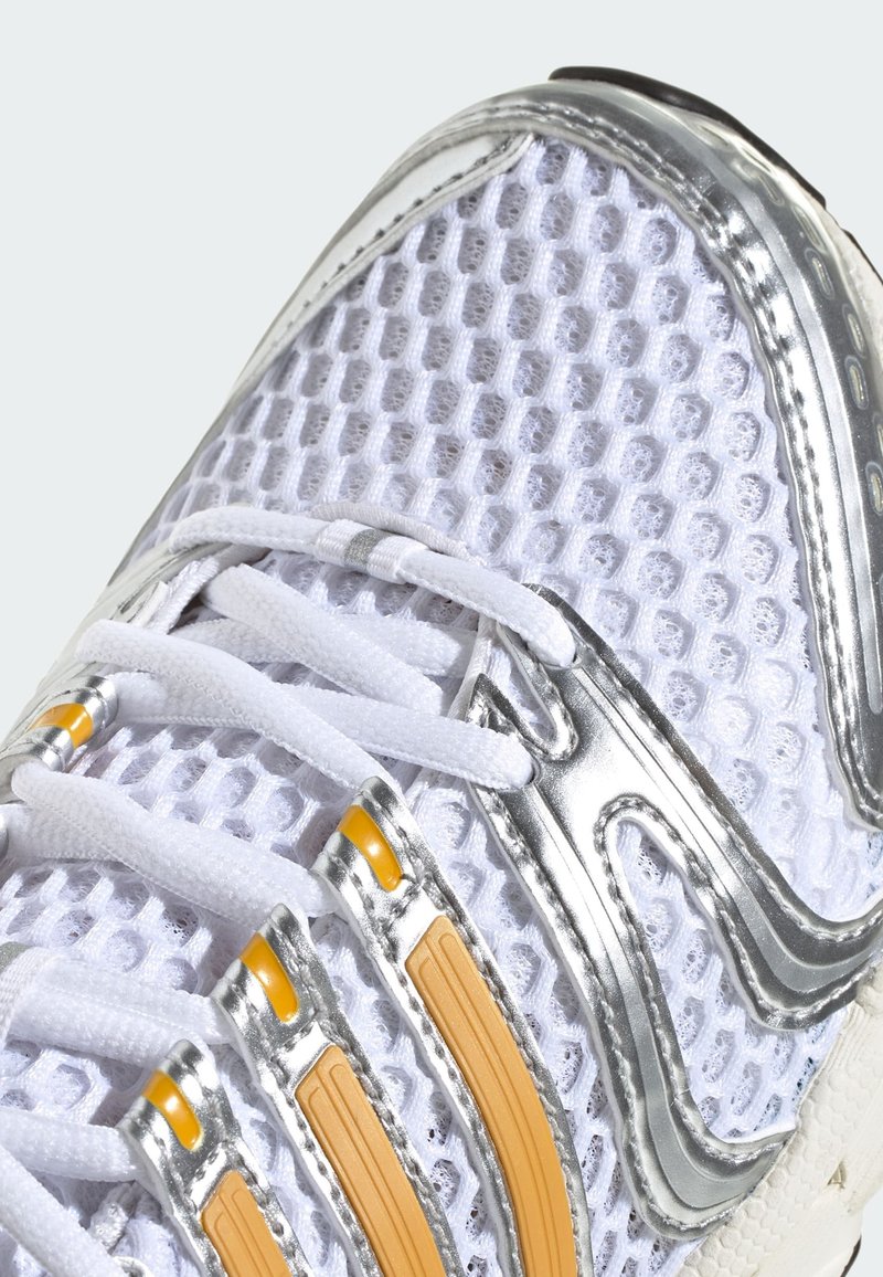 Gros plan sur une chaussure de course en maille blanche avec des accents argentés et orange, dotée de lacets blancs et d'un tissu texturé et respirant.