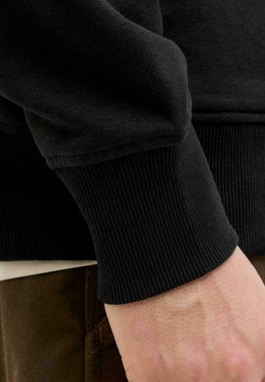 Puño de sudadera negra con textura de canalé, ajustado al cuerpo, mostrando los detalles de la tela. Una mano reposa sobre una camisa de color claro y pantalones marrones.